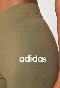Olivgrön sportleggings med en slät textur och vit "adidas"-logotyp tryckt på vänster lår. Hög midja, åtsittande design.