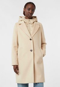 Cappotto in misto lana beige con grandi rever, chiusura a un bottone e due tasche laterali, indossato su una leggera felpa con imbottitura.