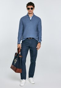 Giovane uomo che indossa un maglione blu con zip e pantaloni scuri, tiene una borsa a tracolla blu navy, con occhiali da sole e sneakers bianche. Sfondo semplice.