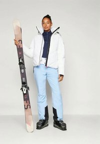 Weißer Pufferjacke mit schwarzen Akzenten, navyfarbige Midlayer, hellblaue Skihose, schwarze Skischuhe; hält einen braunen und schwarzen Ski mit Bindungen.