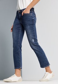 Jeans de mezclilla azul oscuro con un corte ajustado, ligero desgaste en la rodilla y largo de tobillo recortado. Combinados con zapatillas blancas y beige.