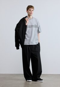 Γκρι μπλουζάκι με μαύρες ρίγες και λογότυπο, συνδυασμένο με φαρδιά μαύρα παντελόνια και μαύρο μπουφάν που είναι περασμένο πάνω από τον έναν ώμο. Άσπρα αθλητικά παπούτσια.