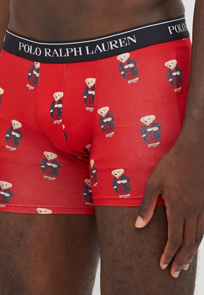Boxers en coton rouge avec un motif répété de nounours portant une robe bleue et tenant une tasse. Ceinture noire avec le logo "POLO RALPH LAUREN".