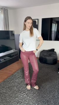 Hvit kortermet skjorte, burgunder tettsittende bukser i kunstlær, brune sandaler, står på teksturert grått teppe med svart bønnepose og TV bak.