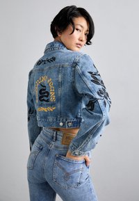 Šviesiai mėlyna apkarpytas džinsinis švarkas su juodais gėlėmis siuvinėtomis detalėmis ir geltonu užrašu. Derinamas su aukštu juosmeniu mėlynais džinsais ir charakteringa etikete.