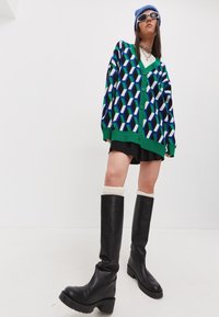 Přesměrně velký zelený cardigan s geometrickým vzorem v modré, černé a bílé barvě. Spárováno s černou sukní a černými koženými botami po kolena.