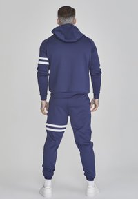 Tengerészkék kapucnis pulóver és hozzá illő joggerek puha anyagból, fehér csíkokkal az ujjakon és a nadrágon, laza szabással és bordázott mandzsettákkal.