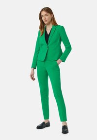 Grüner, maßgeschneiderter Anzug aus Stretchmaterial, bestehend aus einem Ein-Knopf-Blazer, schmal geschnittenen Hosen und schwarzen Loafers. Glatte Textur, lebendige Farbe.