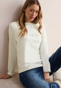 Helle beige Sweatshirt mit "ENDLESS EXCITEMENT" in einer dezenten Schriftart bedruckt, kombiniert mit blauen Jeans, die einen einfachen, entspannten Schnitt bieten.
