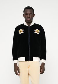 Jaqueta bomber de veludo preta com punhos e gola em branco. Apresenta duas cabeças de tigre bordadas no peito. Fecho frontal com zíper.