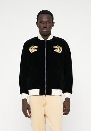 Giacca bomber in velluto nero con polsini e colletto a costine bianchi. Presenta due teste di tigre ricamate sul petto. Chiusura frontale con zip.