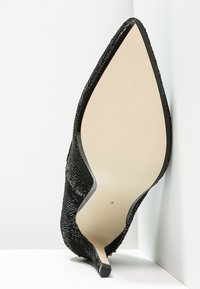 Svart spetsig högklackad sko med texturerad ovandel, beige sula och smal kittenklack som vilar mot vit hörnvägg.