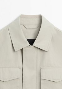 Béžová bunda s límečkem, předním zipem skrytým pod látkovou légou a dvěma náprsními kapsami s klopami a šicím detailem.