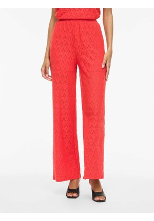 Pantalon rouge à jambes larges avec motif en dentelle, porté avec des talons ouverts noirs, montrant de la taille aux pieds sur fond blanc.