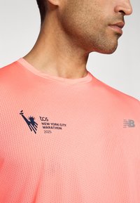 Nahaufnahme einer Person, die ein korallenfarbenes TCS New York City Marathon 2025 Shirt mit einem schwarzen Logo der Freiheitsstatue und einem New Balance Emblem trägt.