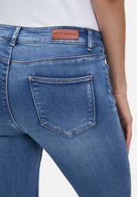 Jean en denim bleu avec une coupe ajustée, une étiquette en cuir marron avec "BETTY BARCLAY" sur la taille, et des poches arrière avec des détails de couture.