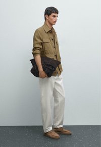 Olijfgroene button-up shirt met borstzakken, witte broek en bruine suède schoenen. Houdt een donkerbruin leren tas vast. Eenvoudig ontwerp.