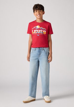 T-shirt rossa in cotone con grafica di montagna, abbinata a jeans in denim blu chiaro a gamba dritta e sneakers bianche con dettagli beige.