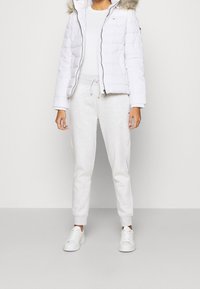 Biała kurtka puchowa z futrzanym kapturem, noszona na białej bluzce i jasnoszarych dresach. Białe sneakersy dopełniają całość.