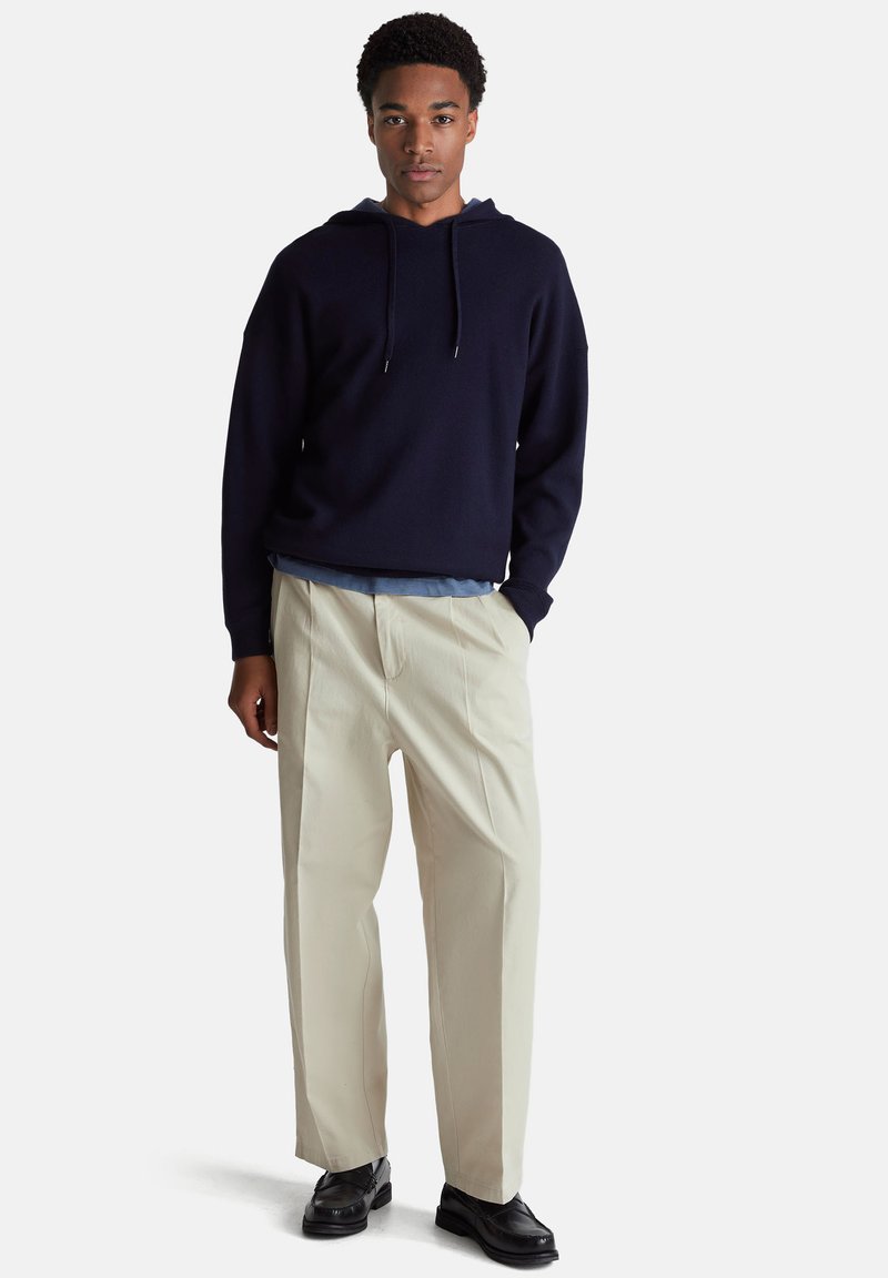 Navy hoody met een ontspannen pasvorm, gecombineerd met crèmekleurige broekspijpen en zwarte schoenen. Gladde textuur, minimalistisch ontwerp en geen zichtbare patronen.