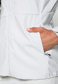 Jasnoszara kurtka zapinana na zamek, z teksturowaną częścią na ramionach, boczną kieszenią oraz gładkim materiałem o subtelnej połysku.