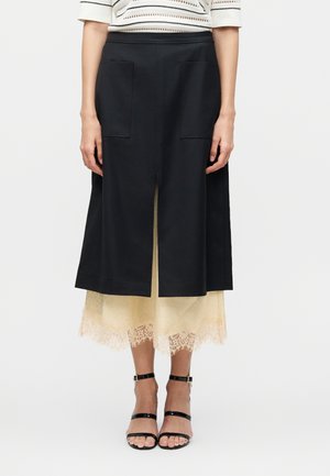 Femme portant une jupe noire mi-longue avec une fente avant et des poches, superposée sur une robe midi beige bordée de dentelle, associée à des talons noirs à bride cheville.