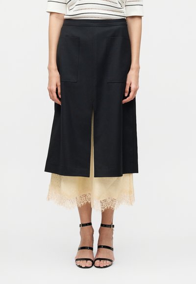 Woman wearing a black knee-length skirt with front slit and pockets layered over a beige lace-trimmed midi dress, paired with black ankle-strap heels.