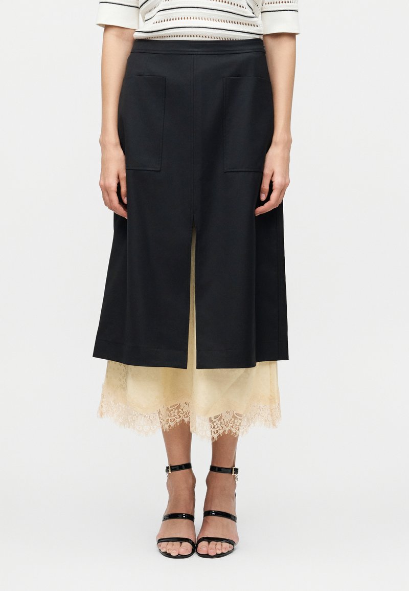 Γυναίκα που φοράει μαύρη φούστα μέχρι το γόνατο με εμπρός σκίσιμο και τσέπες, από πάνω από μπεζ midi φόρεμα με δαντελένια διακόσμηση, σε συνδυασμό με μαύρες γόβες με λουράκι στον αστράγαλο.