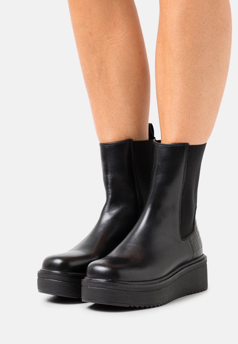 Bottes courtes noires en cuir avec panneaux latéraux élastiques, bout rond et accents texturés sur le talon. Semelle épaisse en caoutchouc. Finition lisse.