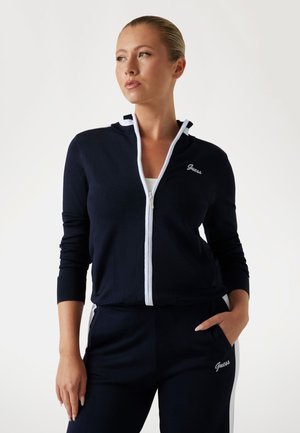 Navy zip-up jacket with white trim, long sleeves, and Guess logo on the chest. The fabric appears soft and textured, designed for comfort.