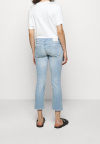 Personne portant un jean court bleu clair, une chemise blanche à manches courtes et des sandales noires ouvertes, debout de dos sur un fond uni.