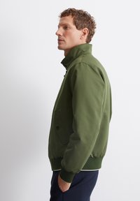 Olive green zip-up jacket with a high collar, side pockets, ribbed cuffs, and hem. Smooth fabric with a structured design.
