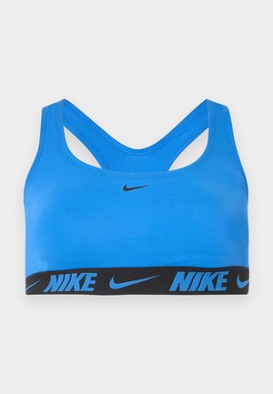 Μπλε αθλητικό σουτιέν Nike με μαύρη ελαστική ταινία που φέρει το λογότυπο και το swoosh της Nike, σχεδιασμός racerback, σε απλό ανοιχτό γκρι φόντο.