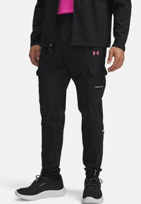 Zwarte cargobroek met elastische boorden, meerdere zakken en een roze Under Armour-logo op de dij. Draagt een paar zwarte sportschoenen.