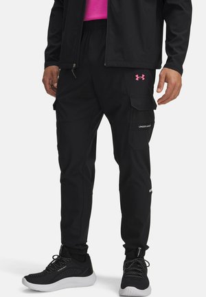 Pantalones cargo negros con puños elásticos, múltiples bolsillos y un logo de Under Armour en color rosa en el muslo. Combinados con zapatillas deportivas negras.