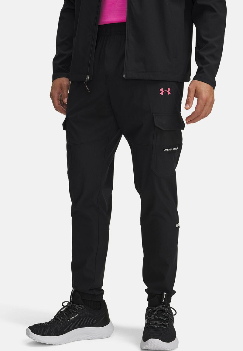 Zwarte cargobroek met elastische boorden, meerdere zakken en een roze Under Armour-logo op de dij. Draagt een paar zwarte sportschoenen.