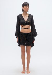 Γυναίκα που στέκεται ξυπόλυτη φορόντας ένα μαύρο δαντελένιο σύνολο δύο τεμαχίων με ιμάντες που τυλίγονται και ένα κίτρινο μπικίνι από μέσα.