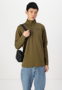 Olivgrüner, langärmeliger Pullover mit halbem Reißverschluss und Logodetail. Kombiniert mit hellblauen, lässig geschnittenen Jeans und einer kleinen schwarzen Umhängetasche.