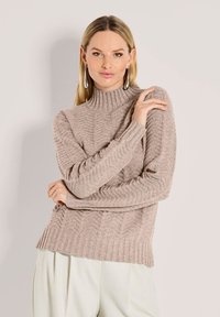 Frau mit langen blonden Haaren, die einen beigen gerippten Rollkragenpullover und weiße Hosen trägt, mit verschränkten Armen vor einem schlichten Hintergrund posiert.