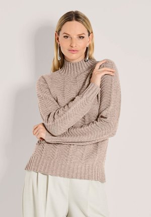 Frau mit langen blonden Haaren, die einen beigen gerippten Rollkragenpullover und weiße Hosen trägt, mit verschränkten Armen vor einem schlichten Hintergrund posiert.