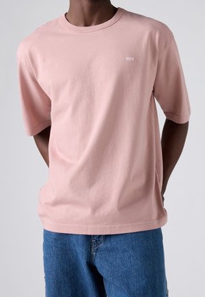 Person iført en lyserød kortærmet t-shirt og blå denimjeans, stående med hænderne bag ryggen.