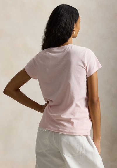 Γυναίκα με μακριά σκούρα μαλλιά, φοράει ανοιχτό ροζ κοντομάνικη μπλούζα και λευκό παντελόνι, στέκεται με το ένα χέρι στη μέση, με την πλάτη γυρισμένη.