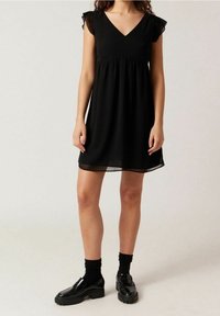 Svart klänning med V-ringning och korta volangärmar. Tillverkad av ett lätt tyg som flödar lätt, med en sammandragning i midjan. Bärs med svarta skor.