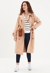 Cappotto beige con bottoni doppi, indossato sopra un maglione a righe blu e bianche, abbinato a jeans scuri e sneakers bianche.