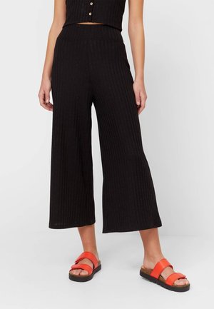 Femme portant un pantalon large noir coupé court, un haut noir sans manches et des sandales orange vif, debout sur fond blanc.