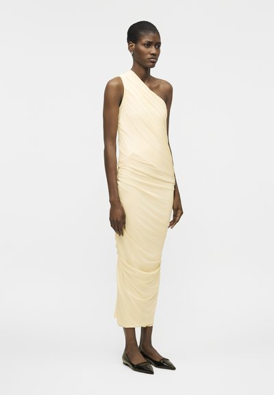 Sieviete stāv, valkājot blāvi dzeltenu vienas pleca ruffled midi kleitu un melnas smailas kurpes, pretējā vienkāršam baltam fonam.