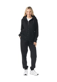 Felpa nera con zip e tasche frontali abbinata a pantaloni da jogging coordinati. Il tessuto ha una consistenza morbida. Sneakers bianche completano l'outfit.