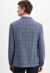 Man met zijn rug naar de camera, gekleed in een blauw geruite blazer met een lichtblauw ruitpatroon en donkere broeken, tegen een witte achtergrond.