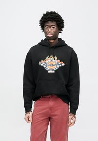 Μαύρο φούτερ με κουκούλα και γραφικό σχέδιο που απεικονίζει φλόγες και ένα ρετρό λογότυπο. Συνδυάζεται με ξεθωριασμένο κόκκινο παντελόνι. Υλικό από βαμβάκι, άνετη εφαρμογή.
