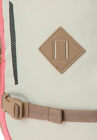 Mochila con un tejido claro y texturizado con detalles en rosa, un parche de goma marrón con el logo y una correa ajustable color beige.
